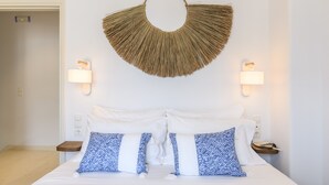 A hotel room with a bed, white bedding, two wall-mounted lamps, and a decorative wall hanging. - Mykonos Perla Town House - Perfect Location with Pool and Private Parking - 2 BR (Mykonos)