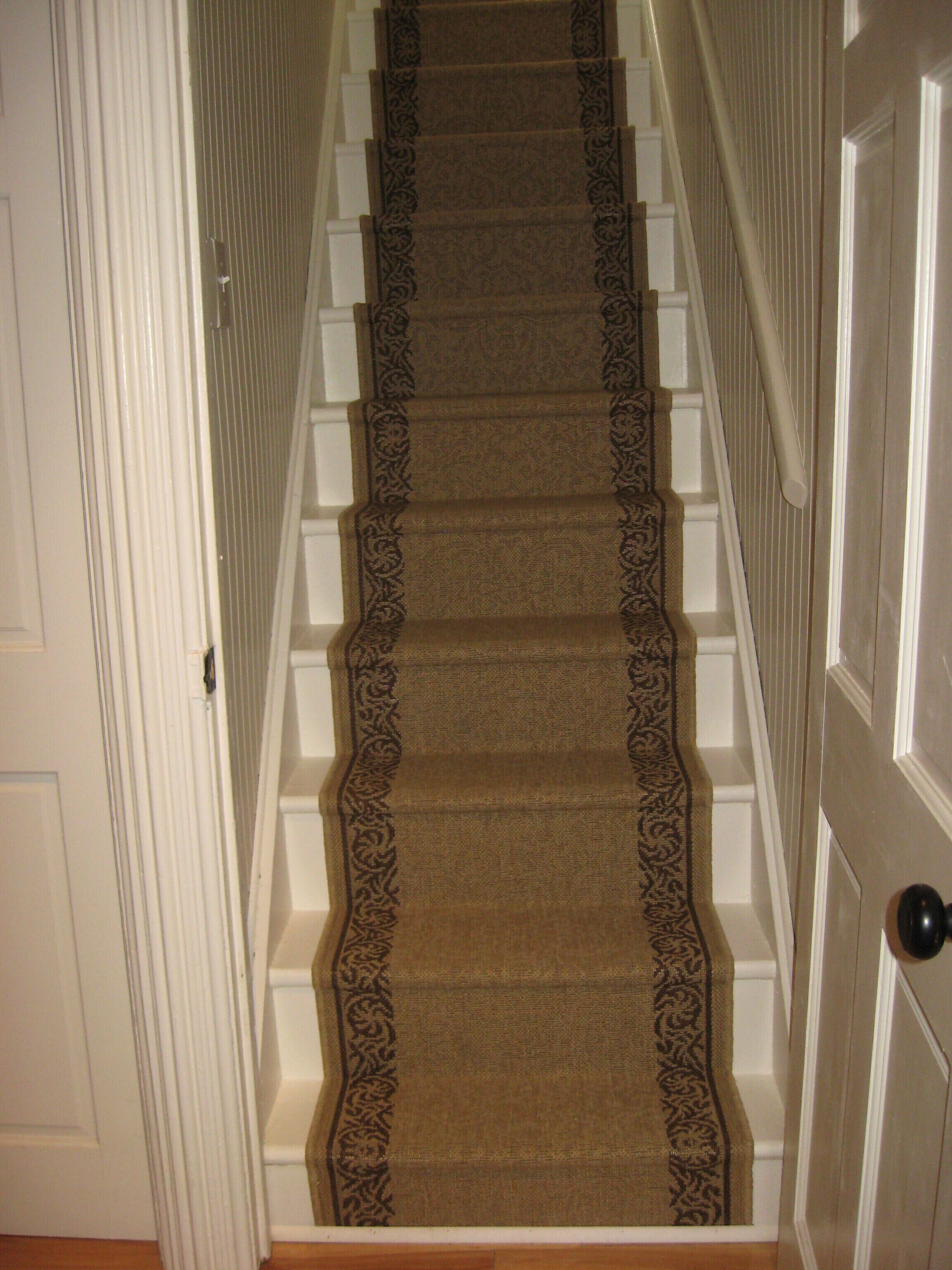 Un escalier recouvert de moquette, avec une bordure à motifs, des murs blancs et une porte dotée d’une poignée de porte noire.