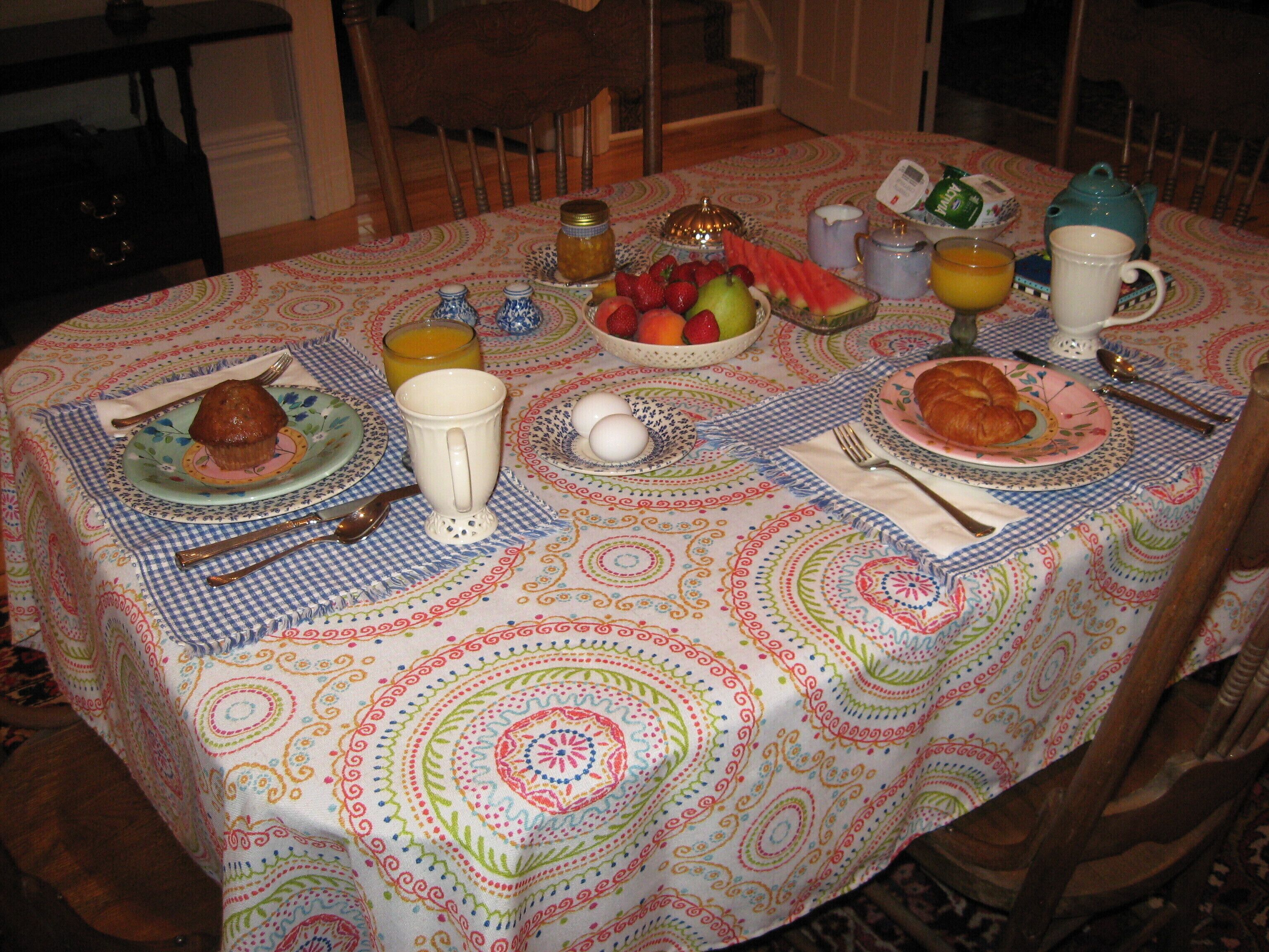 Una mesa con vajilla, cubiertos, una tetera, una jarra y un frutero.