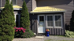 Une maison à deux étages, avec un revêtement extérieur de couleur sombre, un toit jaune et un porche à l’avant avec une porte. On y trouve deux arbres à feuilles persistantes et un parterre de fleurs avec des fleurs roses.