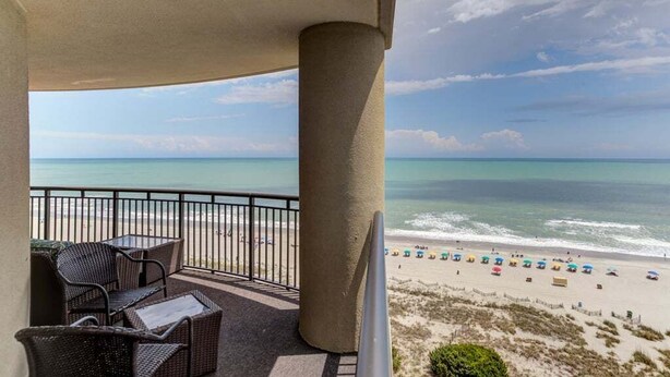Een balkon met rieten meubilair, uitzicht op een strand met parasols en een zeegezicht.