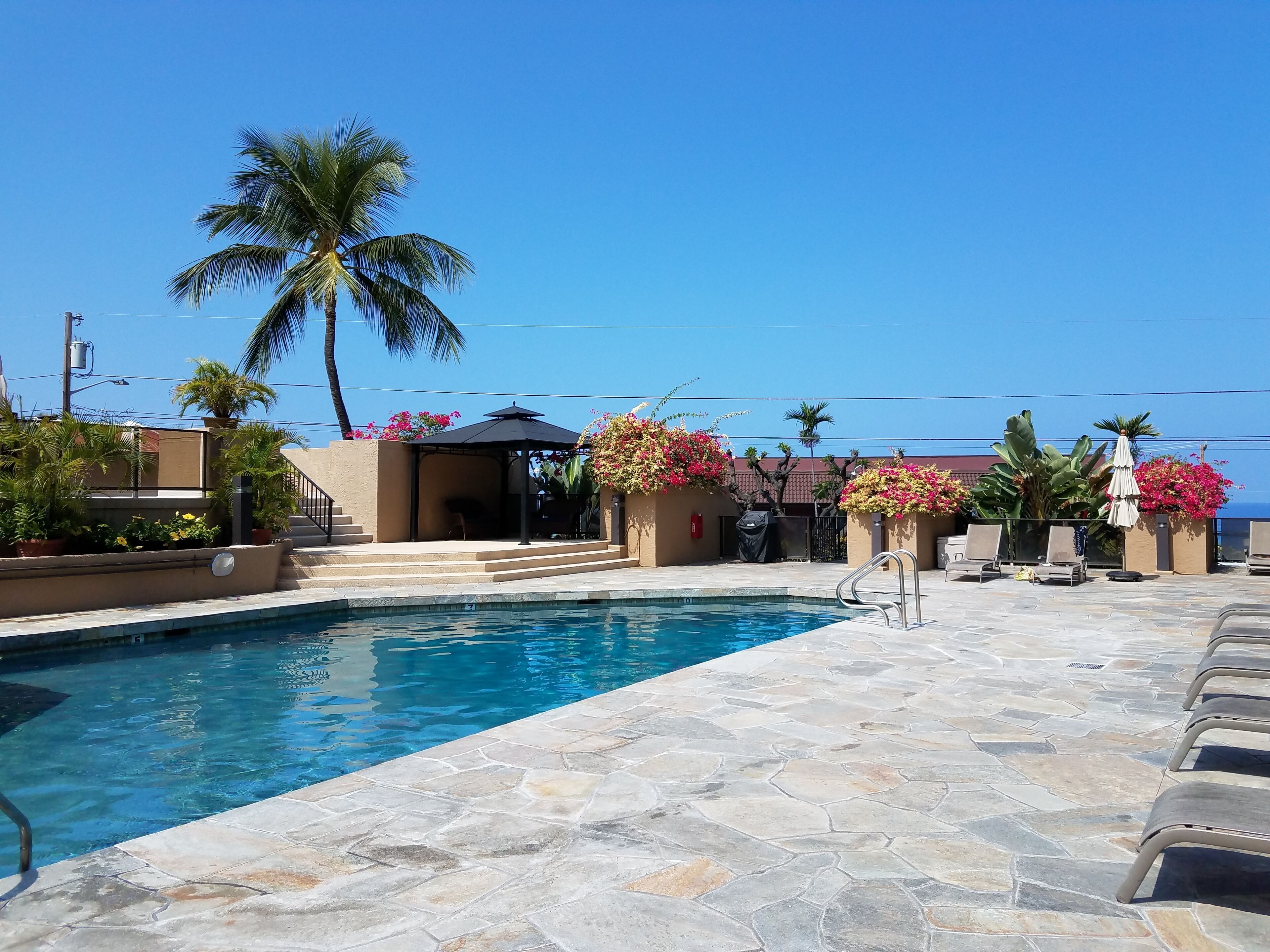 Ein Poolbereich mit klarem, blauem Pool, einer terrassenförmig angelegten, steinverlegten Fläche, Liegestühlen und einer überdachten Lounge mit Sonnenschirmen.