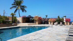 A pool area with a clear blue pool, stone-tiled deck, lounge chairs, and a covered seating area with umbrellas. - Kona Pacific B311 - Quiet, comfortable & walking distance to Town and the beach (Kailua Kona)