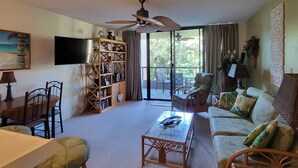 A living room with a ceiling fan, a bookshelf, a television, a dining table with chairs, a sofa, and a coffee table. - Kona Pacific B311 - Quiet, comfortable & walking distance to Town and the beach (Kailua Kona)