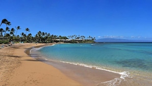 Una playa con aguas turquesas cristalinas, palmeras y una zona de resortes con edificios.