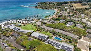 Vista aérea de Napili Shores I2-70, mostrando un entorno costero con playa, océano y una combinación de edificios residenciales y comerciales.