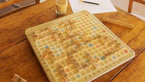 A Scrabble game set on a wooden table with a glass of milk, a pen, and a paper with writing.