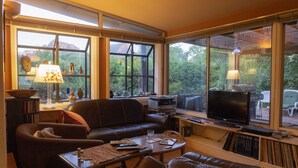 A living room with a brown leather sofa, a coffee table, and a TV. There are windows with a view of trees and mountains.