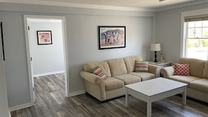 Een woonkamer met een beige bank, een witte salontafel en een ingelijst kunstwerk aan de muur.