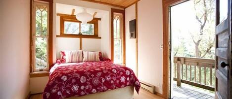 Une chambre avec un plafond en bois, un grand lit recouvert d’une courtepointe à motifs floraux rouges, deux fenêtres et une vue sur une terrasse extérieure.