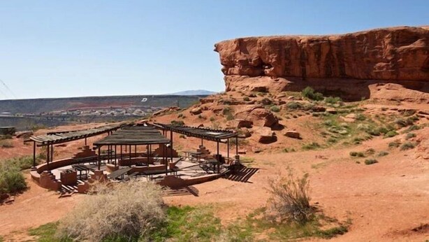 Un área de estar al aire libre rústica con una distribución circular de mesas y sillas, rodeada de vegetación árida natural y una imponente formación rocosa roja al fondo.