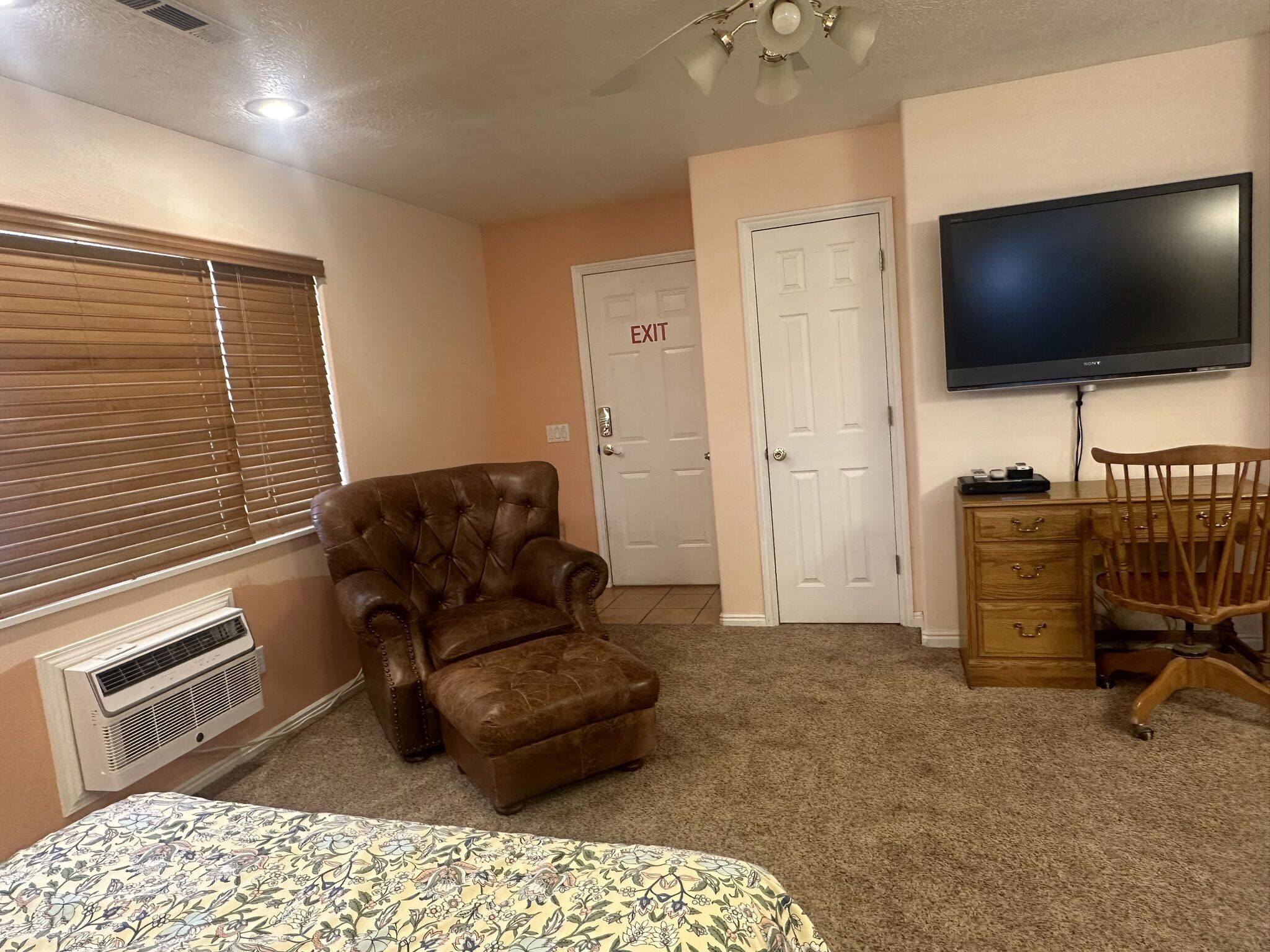 A bedroom with a large bed, a brown leather armchair, a wooden dresser, a television, and an exit door.