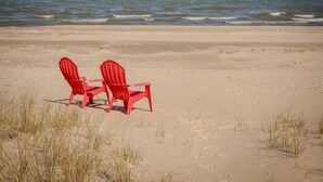 Two red Adirondack chairs on a sandy beach with the ocean in the background.