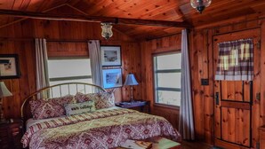 A wooden cabin bedroom with a bed, a nightstand, a lamp, a bench, and a door.