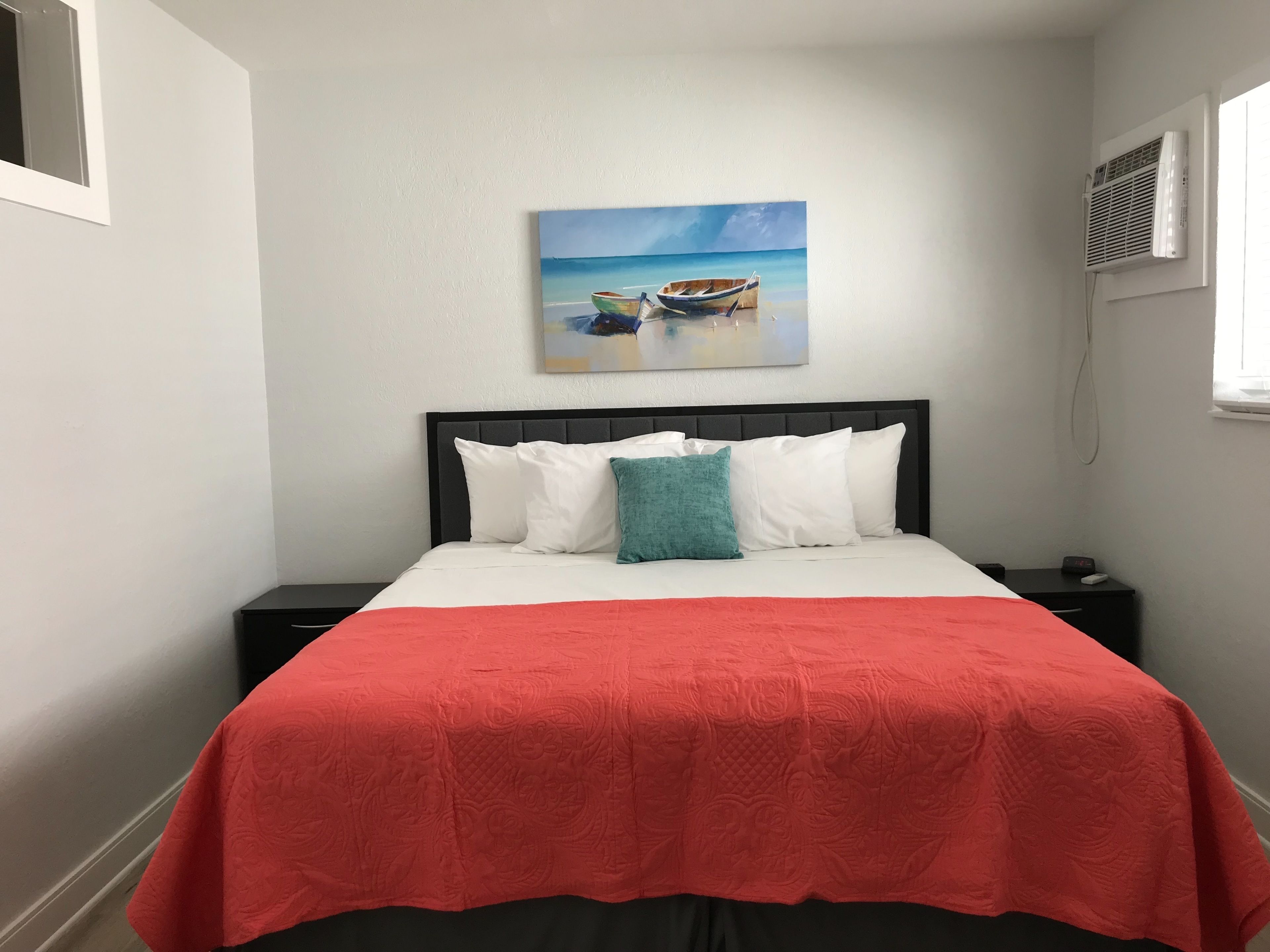 A neatly made bed with a red bedspread and a painting of a beach scene above it.
