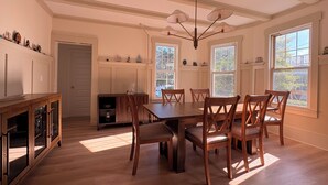 Un comedor con mesa y sillas de madera, una araña y ventanas amplias.