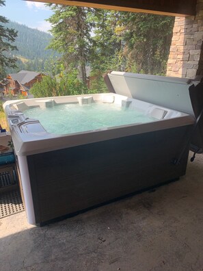 A hot tub with a stone exterior, set outdoors with a view of a forest and houses in the background. - Family Friendly Mountain View Luxury Chalet (Sun Peaks)