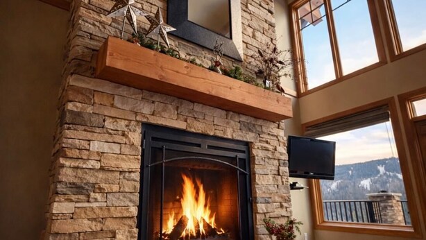 A stone fireplace with a fire, a mounted TV, and a large window with a view.