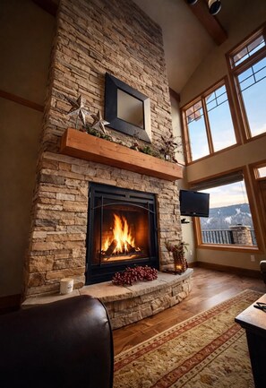 A stone fireplace with a fire, a mounted TV, and a large window with a view. - Family Friendly Mountain View Luxury Chalet (Sun Peaks)