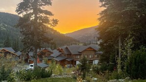 A mountain lodge with multiple wooden cabins, surrounded by lush greenery and tall trees, set against a backdrop of mountains and a sunset sky.