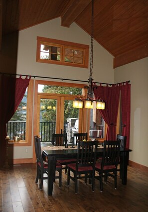 A dining area with a wooden table and chairs, a hanging light fixture, and a view of the outdoors through large windows. - Family Friendly Mountain View Luxury Chalet (Sun Peaks)