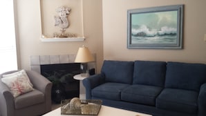 Un salon avec un canapé bleu, un fauteuil gris, un téléviseur, une lampe et un tableau représentant des vagues accroché au mur.
