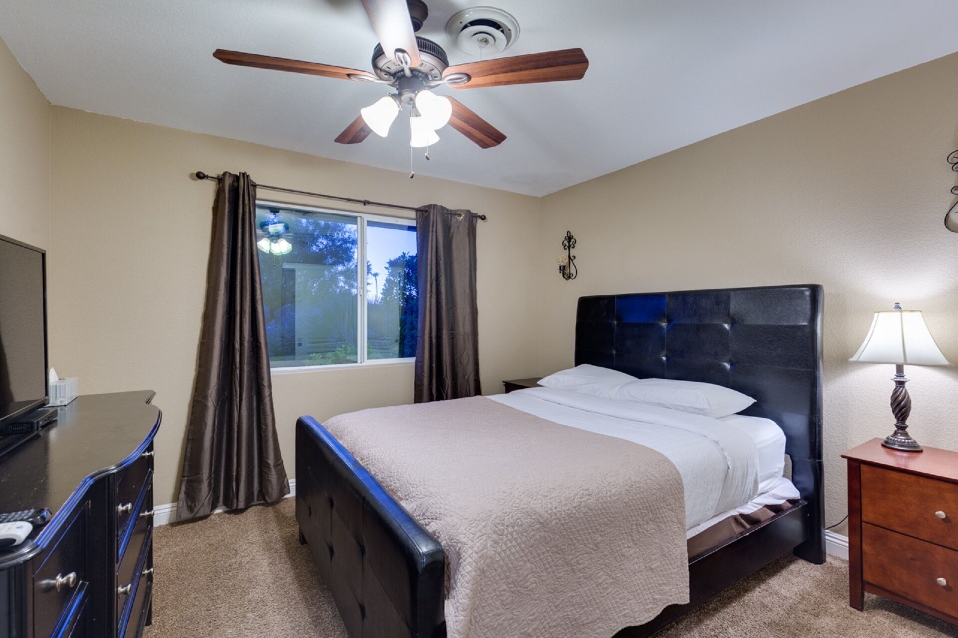Habitación de hotel con cama, escritorio, ventilador de techo, ventana con cortinas y lámpara.