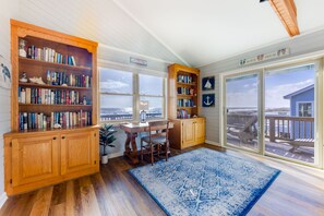 A room with wooden bookshelves, a desk, and chairs, large windows with a view of the beach and ocean, and a blue patterned rug.