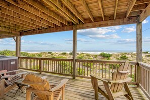A wooden deck with two Adirondack chairs and a hot tub, overlooking a beach with dunes and ocean.