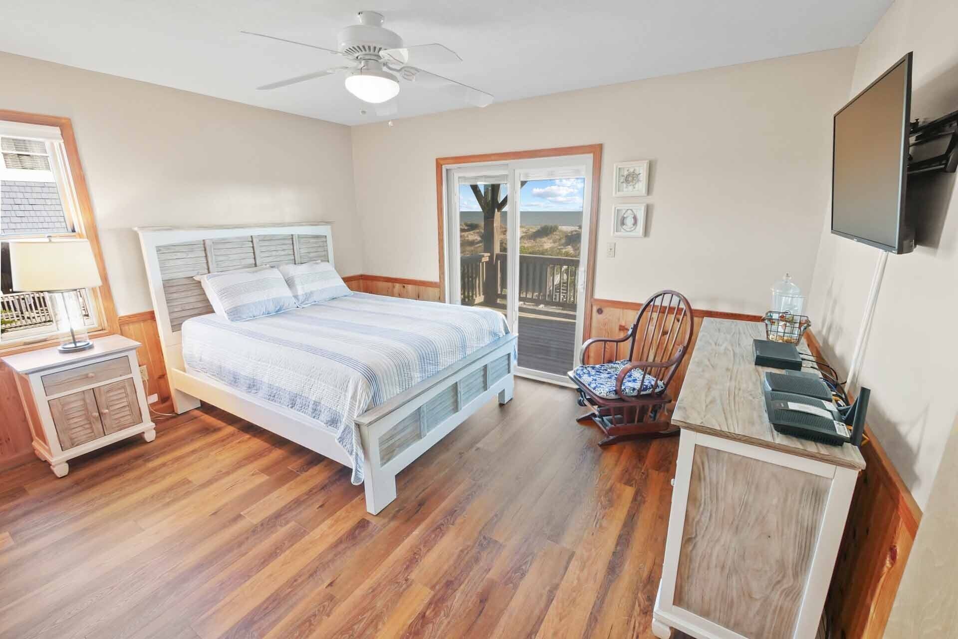 A bedroom with a bed, a chair, a desk, a ceiling fan, and a view through a glass door.