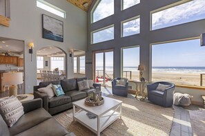 A spacious living room with a large window overlooking the beach, a cozy seating area, and a kitchen in the background. - Big Blue, 13 Steps to the Beach, Guest House, Game Room, Oceanfront, WiFi, Hot Tub (Lincoln County)