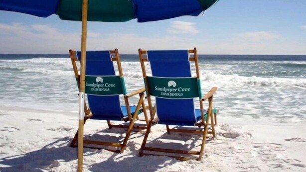 Deux chaises de plage, avec un parasol bleu et vert portant l’inscription « Sandpiper Cove Char Rentals », sont installées sur une plage de sable, avec l’océan en arrière-plan.
