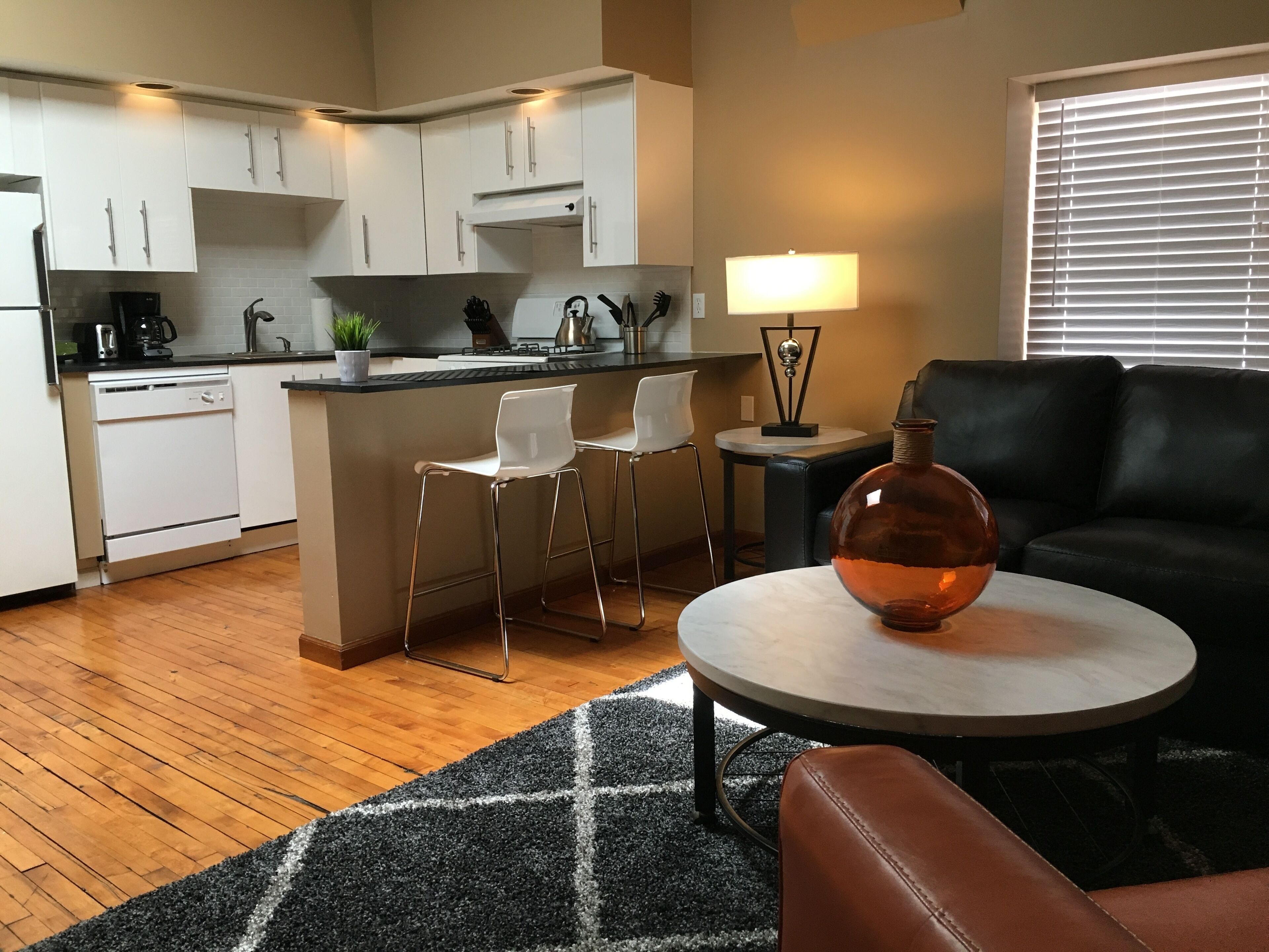 A modern kitchen with white cabinets, a central island with bar stools, a round table with a lamp, and a leather sofa in a living area.