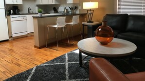 Une cuisine moderne avec des meubles blancs, un îlot central doté de tabourets de bar, une table ronde avec une lampe, et un canapé en cuir dans l’espace salon.