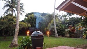 Un barbecue sur une terrasse rouge, avec deux allume-charbon allumés, entouré d’une végétation luxuriante et de palmiers.