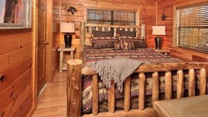 A log cabin bedroom with a wooden bed, two bedside tables, a ceiling fan, and a window with blinds.