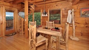A wooden cabin interior with a dining table and chairs, a spiral staircase, and a window with blinds.