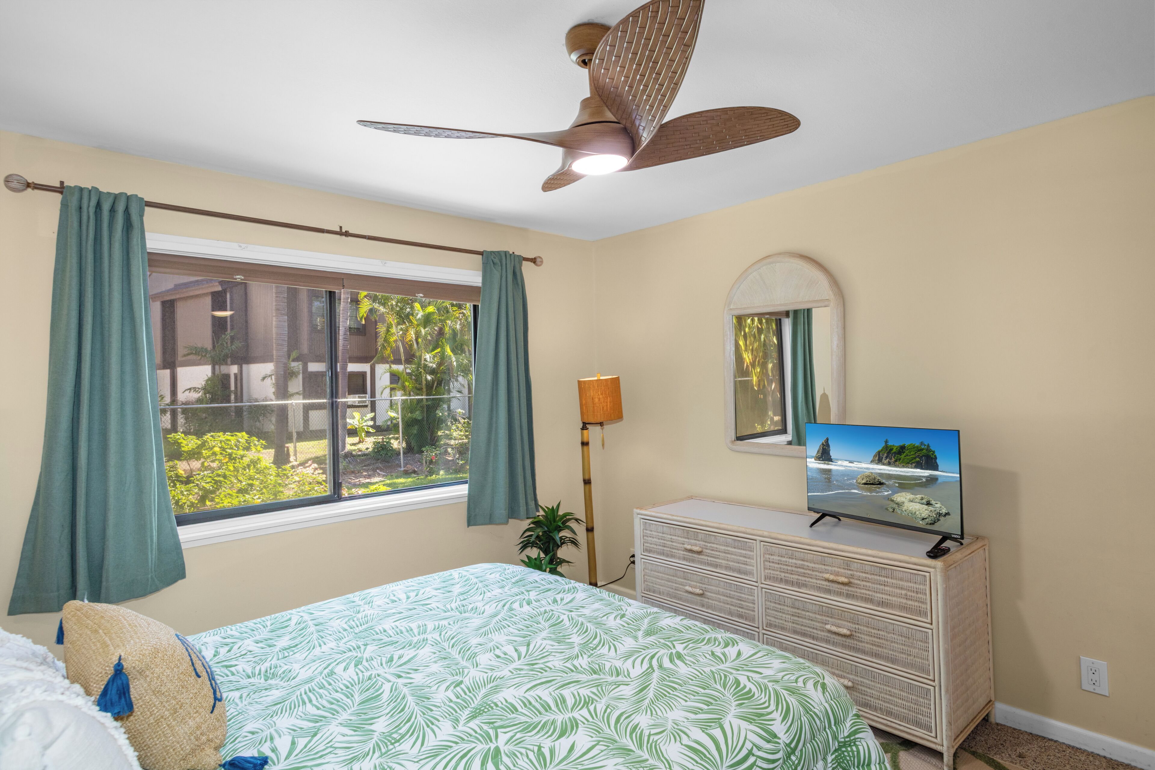A bedroom with a bed, a dresser, a ceiling fan, a window with curtains, and a television.