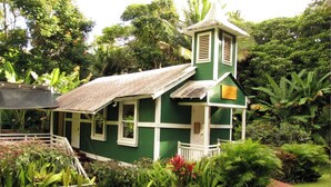 A small, green building with a white porch and a small tower, surrounded by lush tropical vegetation.