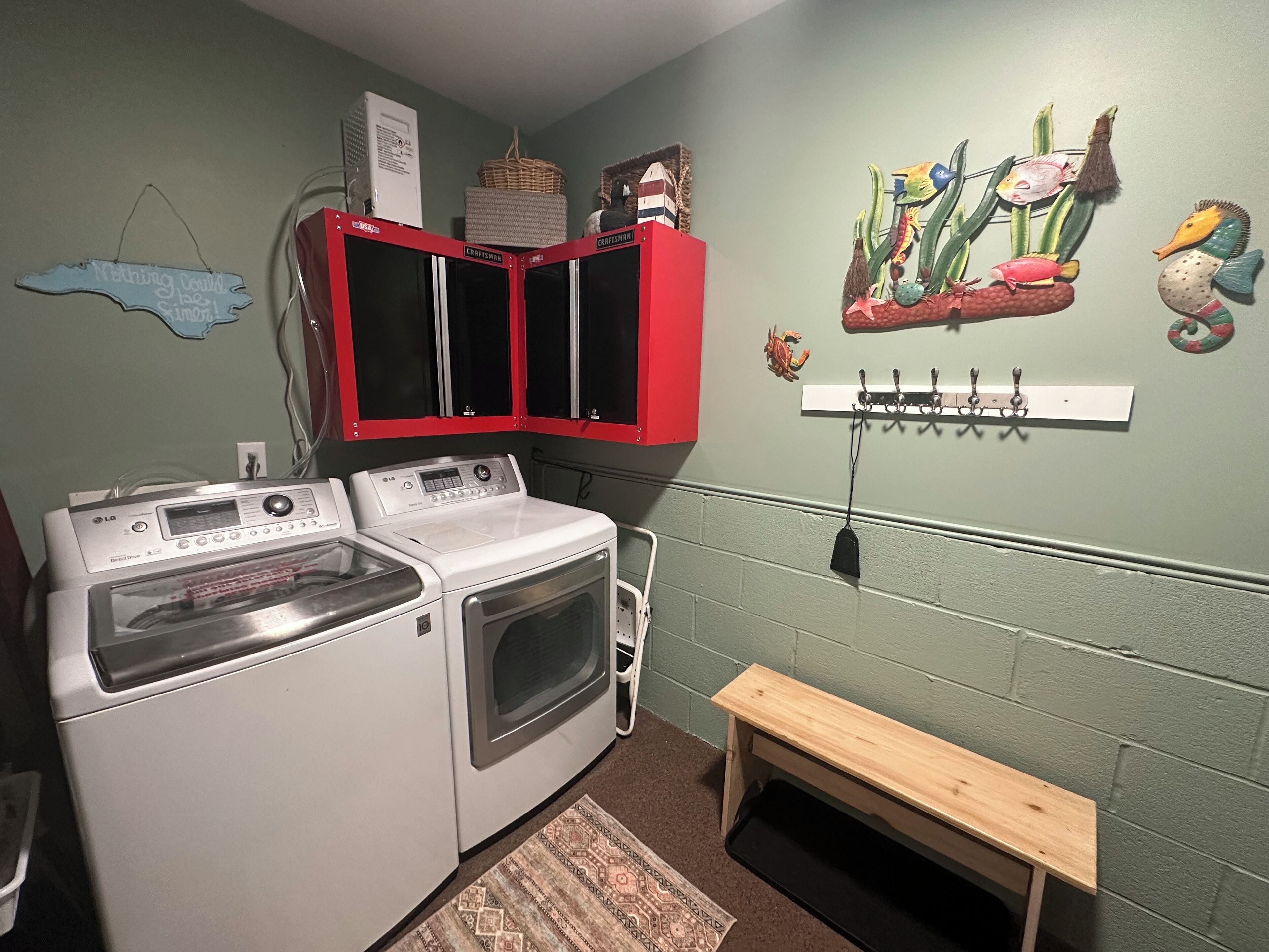 Un espace buanderie comprenant un lave-linge et un sèche-linge à chargement frontal, un meuble rouge, un banc en bois et un étendoir mural orné de motifs représentant des poissons et d’autres créatures marines.