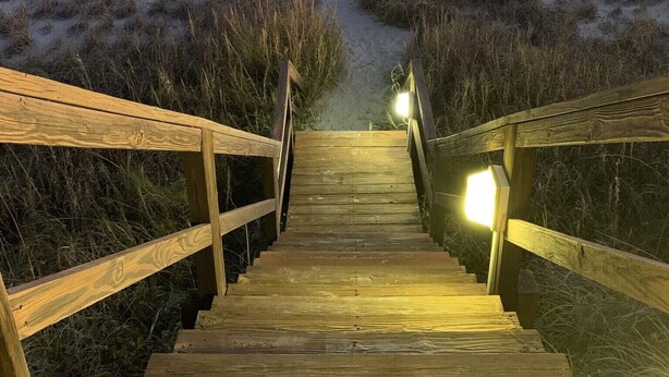 A wooden staircase leading to a beach path with a lifeguard stand visible in the distance.