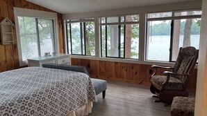 A bedroom with a large bed, a chair, and a view of a lake through multiple windows. - Peaceful Lakefront 3 bedroom Cottage On Quiet Forest Lake  (Gray)