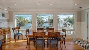 A dining area with a wooden table and chairs, large windows with a view of trees and a white fence, and a checkered rug on the floor. - Rehoboth Beach Lake Front Private Pool Home Sleeps 12 (Rehoboth Beach)