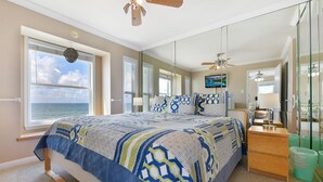 A bedroom with a large bed, a wooden nightstand, a mirror, and a view of the ocean.
