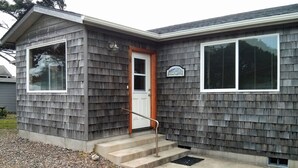 A single-story building with a gray shingle exterior, a white door, and a small porch with steps leading to the entrance. - Single level home, near beach. Ultra clean. Great location -Walk to everything! (Yachats)