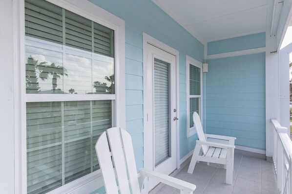 A covered porch with two white Adirondack chairs, a door, and a window with blinds.
