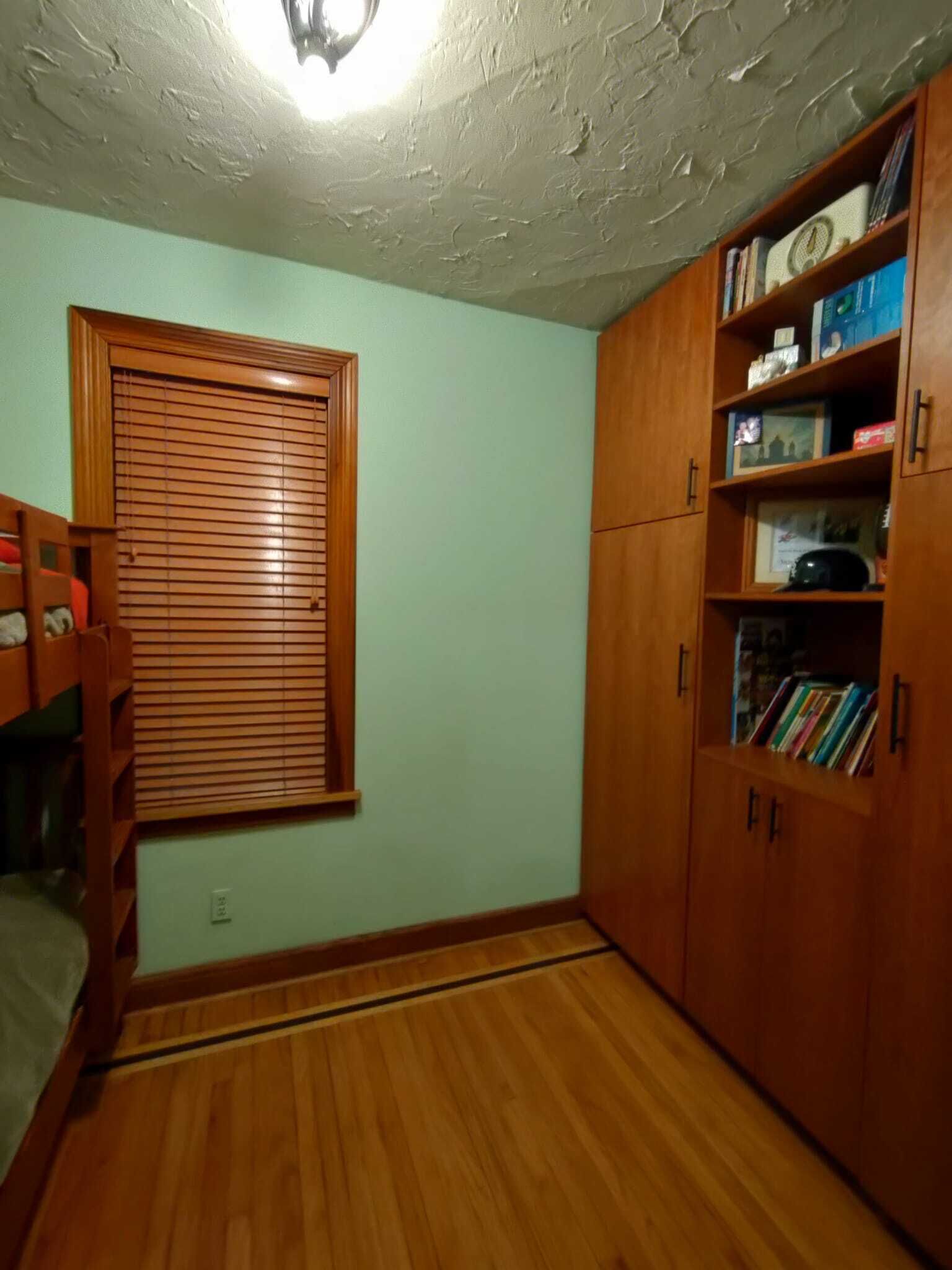 A room with a bunk bed, wooden flooring, a window with blinds, and a bookshelf filled with books and decorative items.