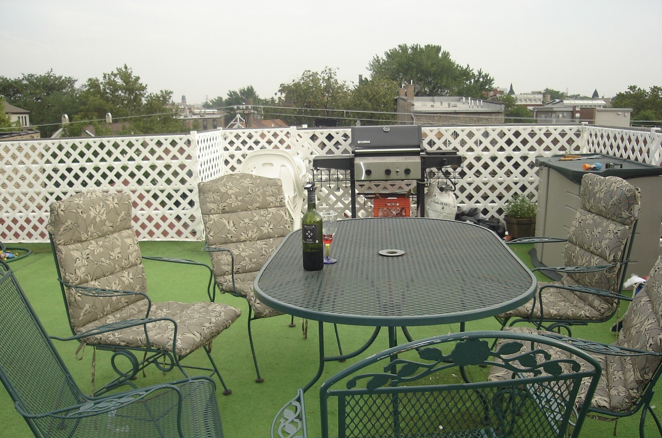 A rooftop patio with a round metal table and four patterned chairs, a grill, and a small potted plant.