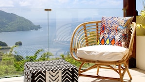 Une chaise en osier agrémentée d’un coussin coloré et d’un pouf noir et blanc à motifs, sur un balcon doté d’une rambarde en verre, d’où l’on peut admirer un plan d’eau et la verdure environnante.