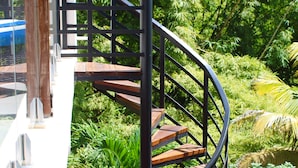 Un escalier en colimaçon avec des rampes en métal et des marches en bois, entouré d’une végétation luxuriante.